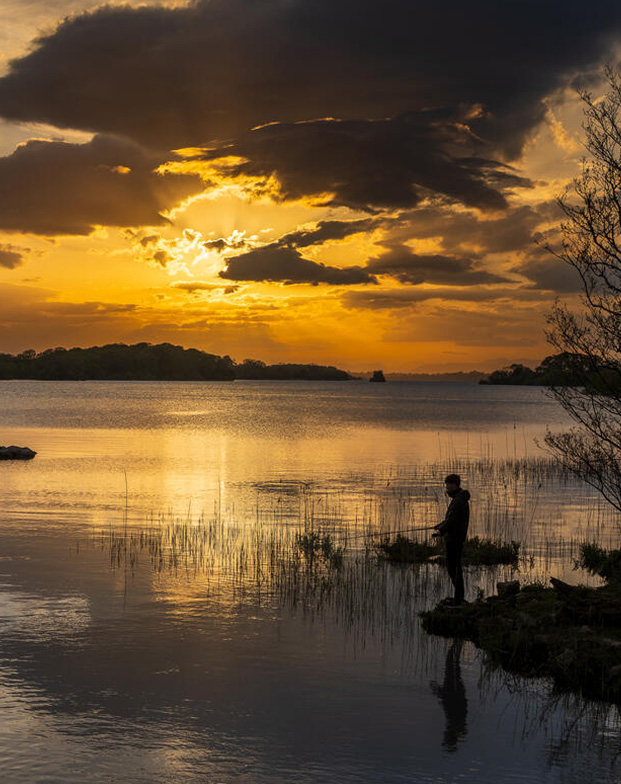 Sunset silhouette of person fishing at the edge of a lake